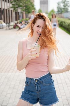 Young beautiful red-haired woman with braces drinks cooling cocktail outdoors in summer. Portrait of a smiling girl with freckles.