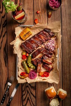 Grilled pork ribs served with grilled corn, salat, bbq sauce, salt pepper and cucumber on parchment paper on a wooden table. Top View. Still life. Flat lay.