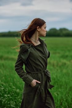 a beautiful red-haired woman stands in a field in a long dark raincoat, holding her hands in her pockets. High quality photo