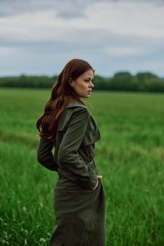 a beautiful red-haired woman stands in a field in a long dark raincoat, holding her hands in her pockets. High quality photo