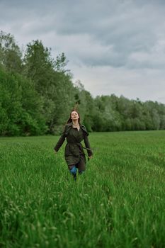 a beautiful woman in a long raincoat runs across a field in high grass in spring in cloudy weather. High quality photo