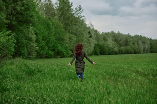 a woman in a long raincoat runs across a field in tall green grass in cloudy weather in spring. High quality photo