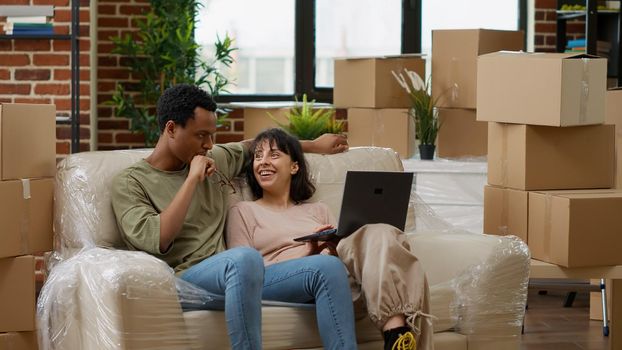 Diverse couple browsing internet to buy decor on laptop, buying home furniture on online shopping to decorate living room apartment. Moving in rented household property together. Tripod shot.