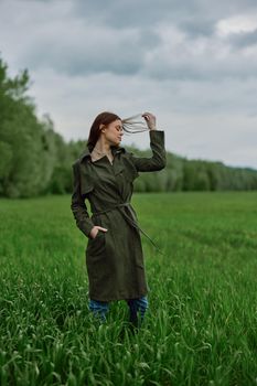 a beautiful woman in a dark coat stands in a green field in the spring in rainy weather and straightens her hair. High quality photo