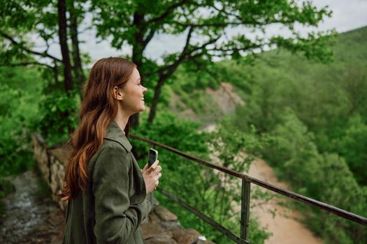 a woman in a raincoat takes pictures on the phone of a beautiful view of the forest. Travel, technology, mobile photography. High quality photo