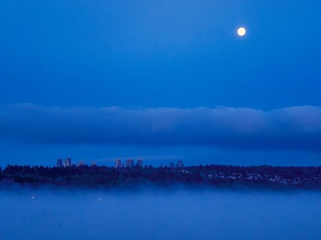 Night city in clouds with full moon over in the sky.