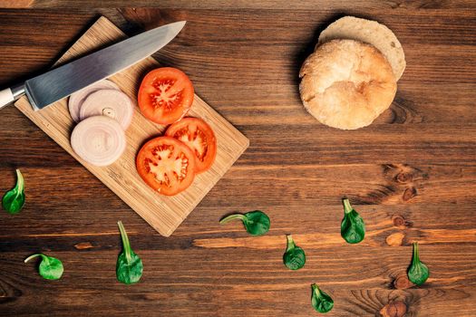 knife with slices of tomato and onion on a cutting board next to a loaf of bread and some green leaves on a wooden table, top view, vegan food and healthy lifestyle concept