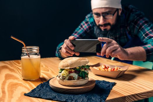chef taking a photo with the phone of his vegan burger with lettuce and sauce, fries with ketchup and healthy drink on a wooden table, technology and gourmet lifestyle concept, selective focus