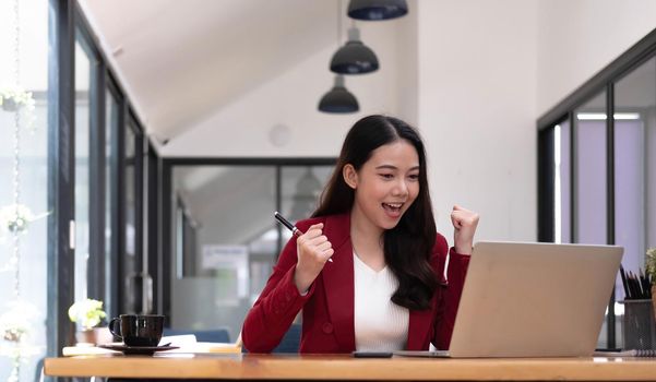 Excited happy woman looking at the laptop celebrating an online win, overjoyed young asian female screaming with joy at office