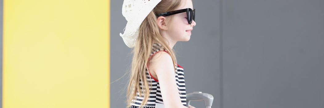 Portrait of well dressed little girl going on vacation carry yellow suitcase with her. Kid in cute hat and lovely summer dress. Airport, childhood concept