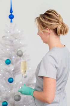 Beautiful young caucasian smiling female doctor in protective gloves with glass of champagne standing near the christmas tree