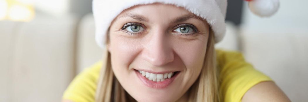 Portrait of beautiful blonde young woman posing on camera in santa claus hat and smile. Celebrating new year alone at home. Holiday, christmas mood concept