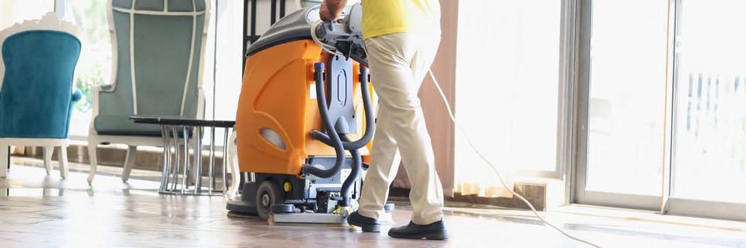 Close-up of male cleaner cleaning interior of modern hotel lobby. Man in uniform with floor washer machine. Janitor in hall using washing vacuum cleaner. Cleaning service concept
