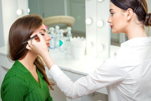 Makeup artist applies eyeshadow powder on eyes of young caucasian woman by brush tool in beauty salon