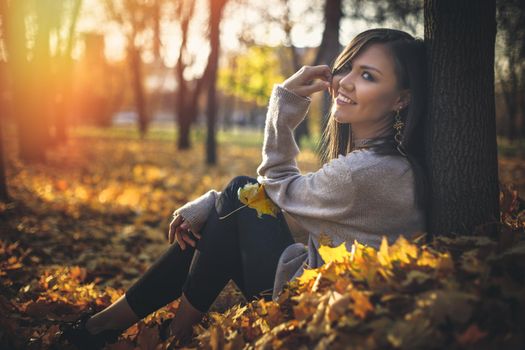 Young happy mixed race woman sitting near tree in autumn park at sunset looking at camera.