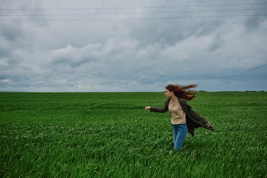 a woman in a coat walks through a green field in rainy weather. Strong wind, harmony with nature. High quality photo