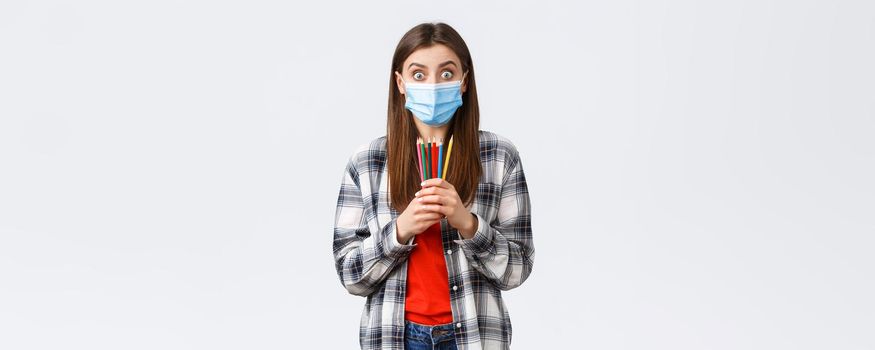 Social distancing, leisure and hobbies on covid-19 outbreak, coronavirus concept. Enthusiastic cute girl in medical mask trying new thing on self-quarantine, showing colored pencils, learn how draw.