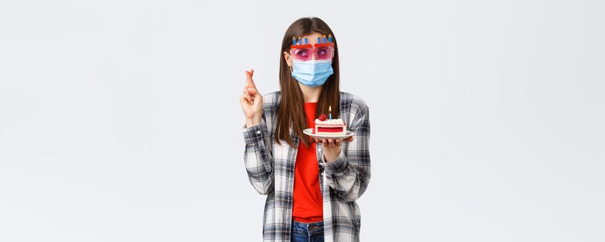 Coronavirus outbreak, lifestyle on social distancing and holidays celebration concept. Excited cute birthday girl in glasses and medical mask, cross fingers making wish as hold b-day cake with candle.