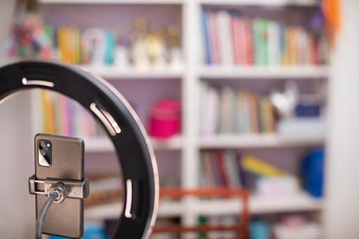 Cell phone mounted in the holder. A circular lamp to illuminate the face. In the background in a blur a bookcase with many colored books
