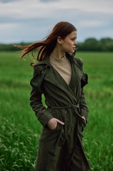 a beautiful red-haired woman stands in a field in a long dark raincoat, holding her hands in her pockets. High quality photo