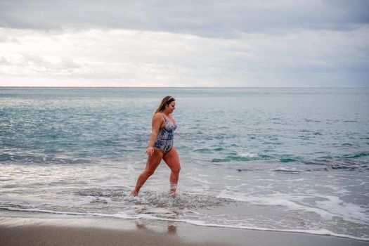 A plump woman in a bathing suit enters the water during the surf. Alone on the beach, Gray sky in the clouds, swimming in winter