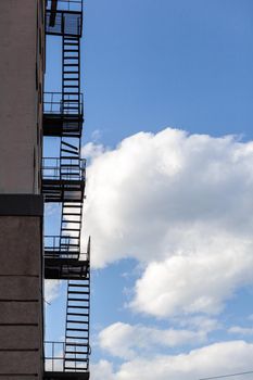 Silhouette of a fire escape on a high-rise building against a blue sky with clouds. Some of the stairs are broken. There is free space for text