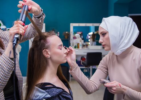 Two craftsmen curl the girl's hair and paint her eyes with a brush. Make-up artist and hairdresser create an image of a young woman. Business concept - beauty salon, facial skin and hair care.