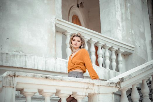 A middle-aged woman looks like a good blonde with curly beautiful hair and makeup on the background of the building. She is wearing a yellow sweater