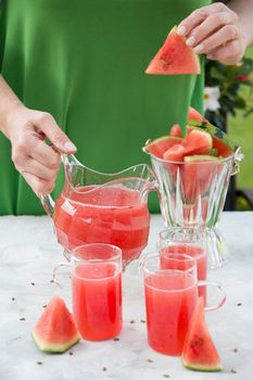 a woman in a green dress pours a red refreshing drink from the pulp of a watermelon into a glass glass, bright natural background