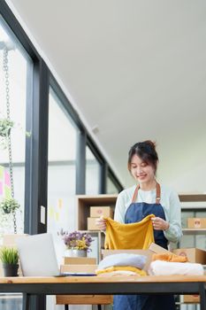 Portrait of a small startup Asian female entrepreneur SME owner picking up a yellow shirt before packing it in an inner box with a customer. Online Business Ideas and Freelance.