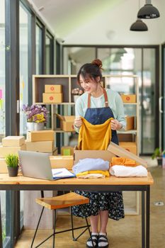 Portrait of a small startup Asian female entrepreneur SME owner picking up a yellow shirt before packing it in an inner box with a customer. Online Business Ideas and Freelance.