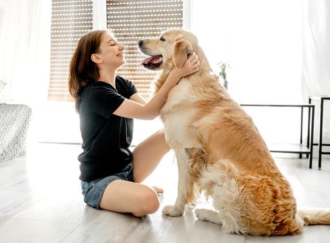 Pretty girl with golden retriever dog sitting on floor at home and petting pet friend. Beautiful female teenager person with purebred doggy