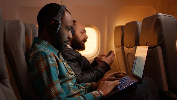 African american freelancer using laptop on flight, travelling on work trip during sunset in economy class. Working on computer and flying on airplane, aviation journey.