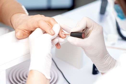 Manicure master in protective gloves applying beige nail polish on female nails in beauty salon