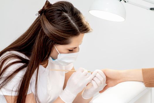 Manicure master with protective mask in rubber gloves applying beige nail polish on female nails in beauty salon