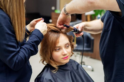 Professional hairdresser twists curls of long light brown hair of woman with curling iron in beauty salon. Hairdressing procedures