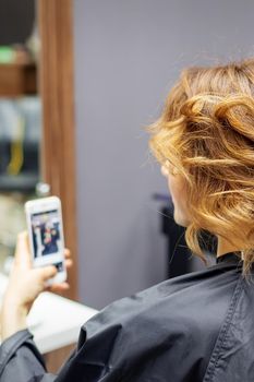 Hairdresser doing hairstyle for young woman with red curly hair and with smartphone in her hands in beauty salon