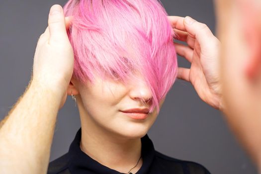 Hairdresser checking short pink hairstyle of young woman on gray background