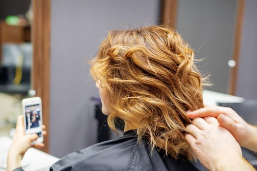 Hairdresser doing hairstyle for young woman with red curly hair and with smartphone in her hands in beauty salon