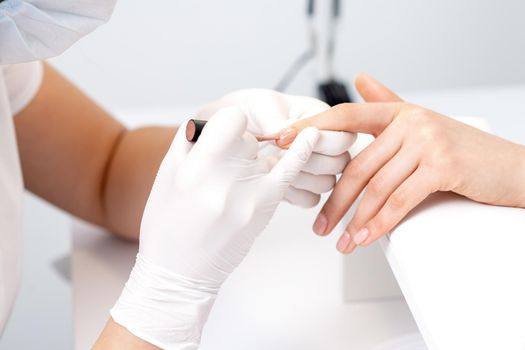 Manicure master in protective gloves applying beige nail polish on female nails in beauty salon