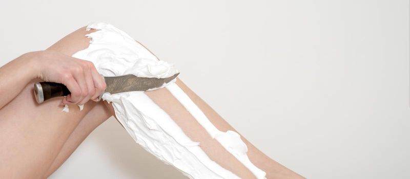 Female beautiful legs of young woman with foam during shaving by knife on white background