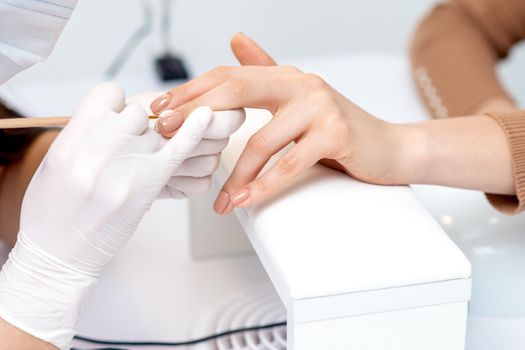 Manicure master in protective gloves applying beige nail polish on female nails in beauty salon
