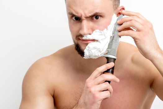 Young caucasian man shaving beard with knife on white background