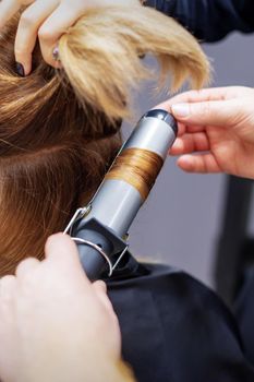 Professional hairdresser twists curls of long light brown hair of woman with curling iron in beauty salon. Hairdressing procedures