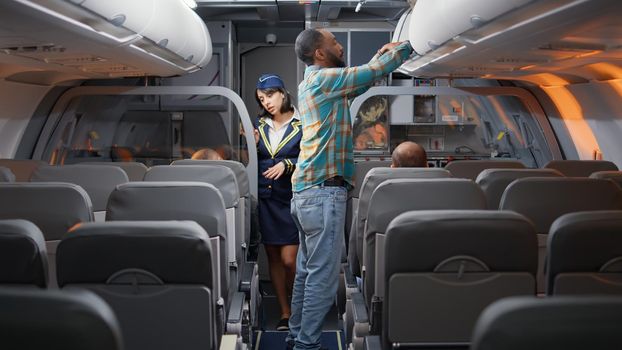 Diverse passengers boarding on airplane to fly in economy class, being seated by flight attendant to travel on vacation trip. Talking to stewardess in plane, flying with airline crew.