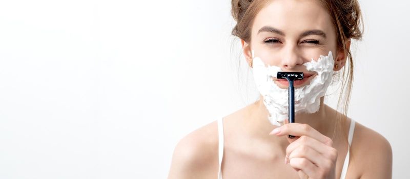 Beautiful young caucasian woman shaving her face by razor on white background. Pretty woman with shaving foam on her face