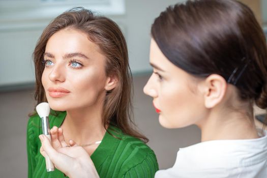 Make up artist applying professional make up of tonal foundation on the face of beautiful young caucasian woman in make up room. Base for make up.