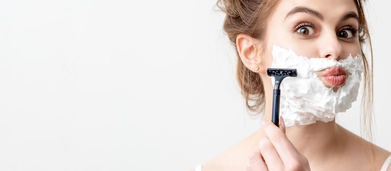 Beautiful young caucasian woman shaving her face by razor on white background. Pretty woman with shaving foam on her face