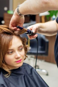 Professional hairdresser twists curls of long light brown hair of woman with curling iron in beauty salon. Hairdressing procedures