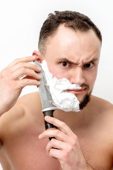 Young caucasian man shaving beard with knife on white background